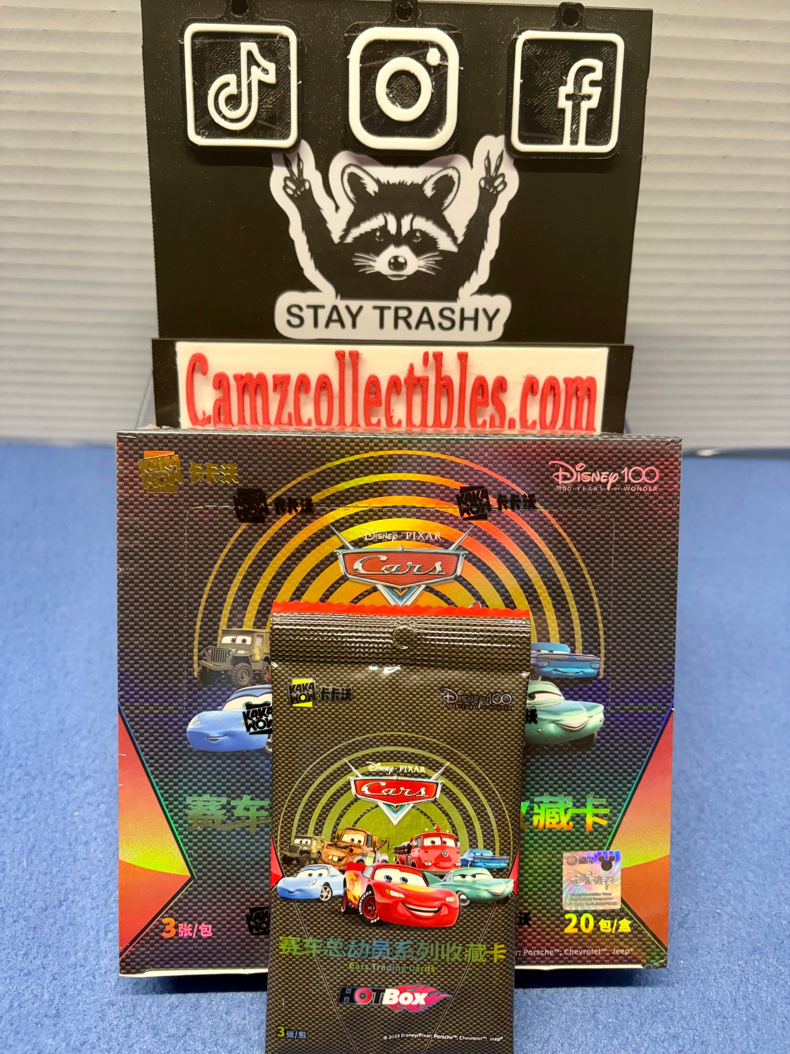 Disney Cars collectible card pack set displayed on a blue surface, featuring a main illustrated card box, a sealed foil pack, and a black sign behind it with social media icons and the text 'STAY TRASHY' and 'Camzcollectibles.com'.