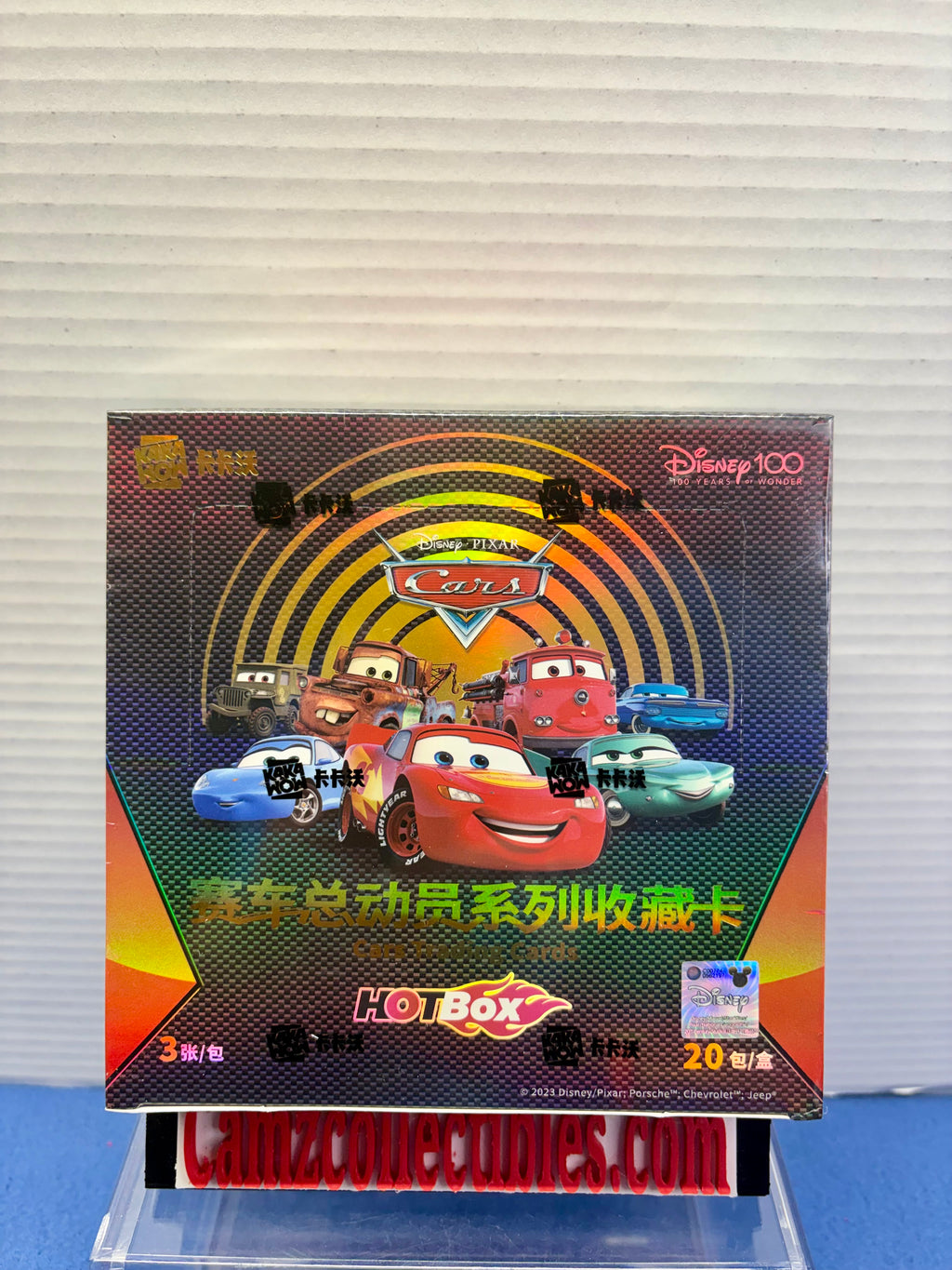 A Disney Cars Hot Box collectible card pack box displayed on a clear stand against a white ribbed wall background. The packaging features Lightning McQueen and other Disney Cars characters with Chinese text.